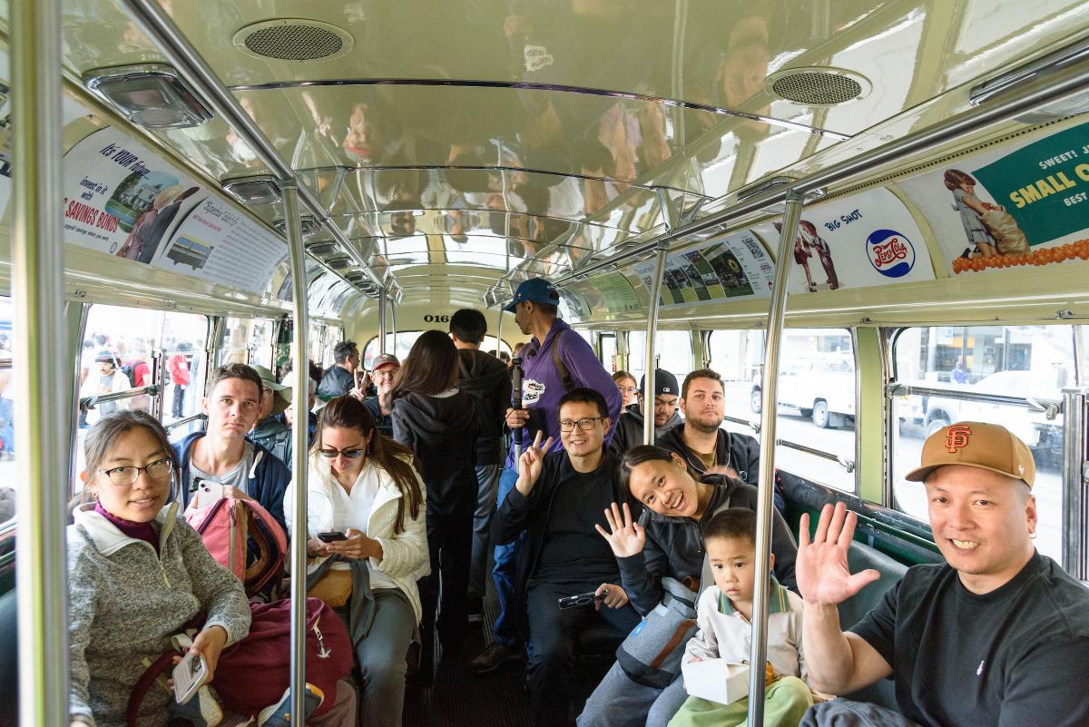 A group prepares to ride a vintage bus during Muni Heritage Weekend. A group of people sit in a vintage bus. A few of them wave to the camera.
