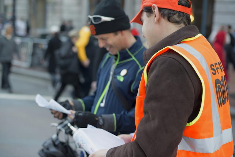 Muni ambassador in orange safety vest assists bicyclist during Super Bowl City event.