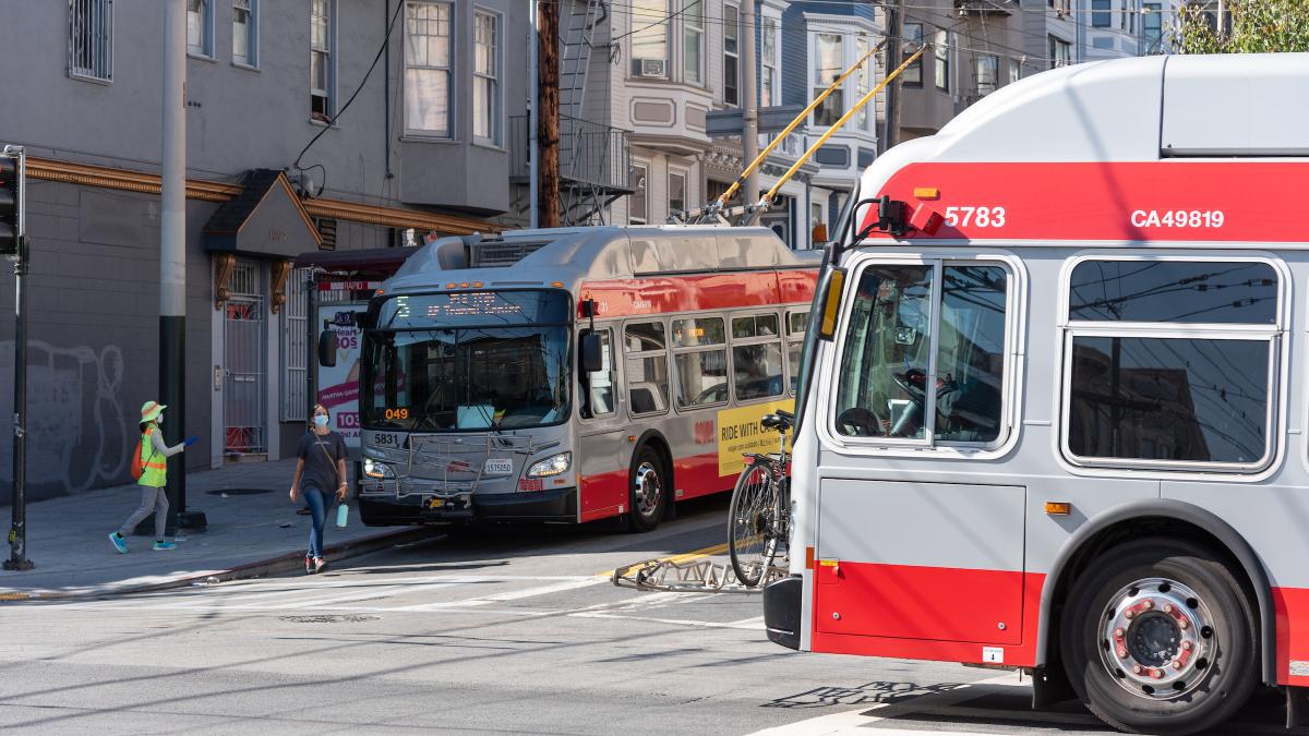 Image of a 5 Fulton bus stopped at an intersection where one rider waits to board as another exits the bus. Another bus passes through the same intersection.