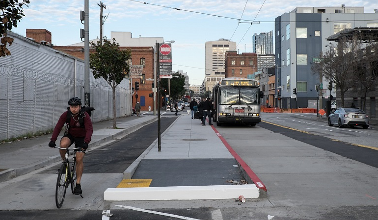 A man rides in a bike lane between the sidewalk and a concrete island where people board a Muni bus on 11th Street at Harrison Street.