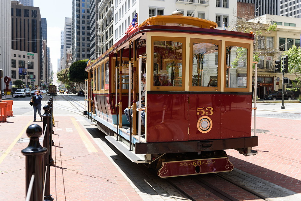 Cable Car 53 paused on California Street. The car is red and gold with fresh paint. A woman walks by.