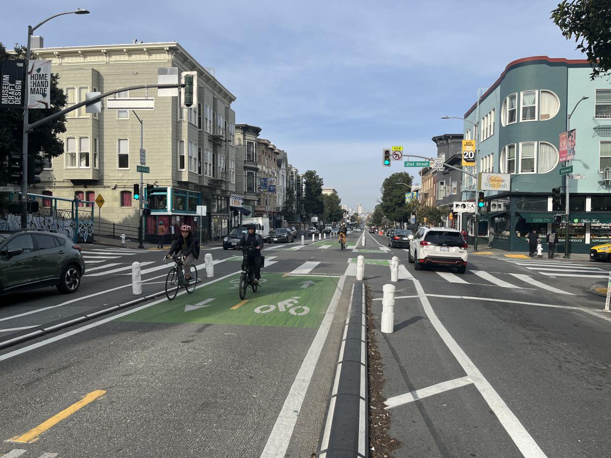 Two people ride bikes on a green, center-running bike lane on Valencia. A white SUV drives in the vehicle lane on the right side.