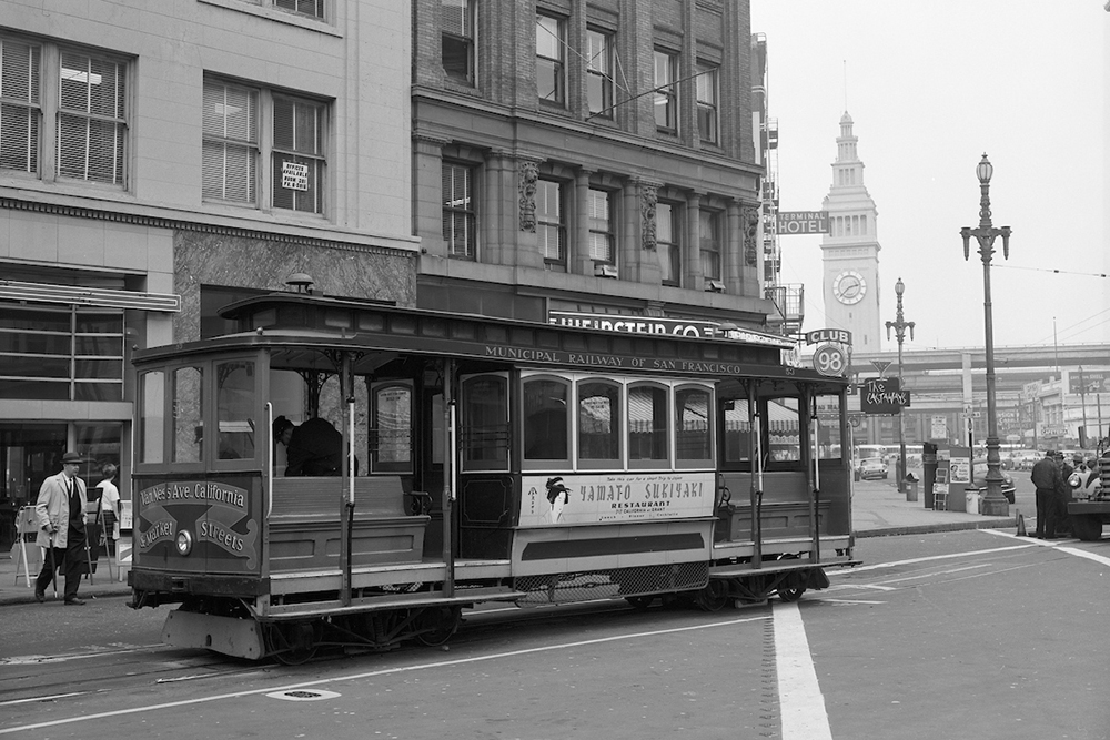 Black and white photo featuring Cable Car 53 moving on Market Street. People walk in the background, where we also see the Ferry Building.