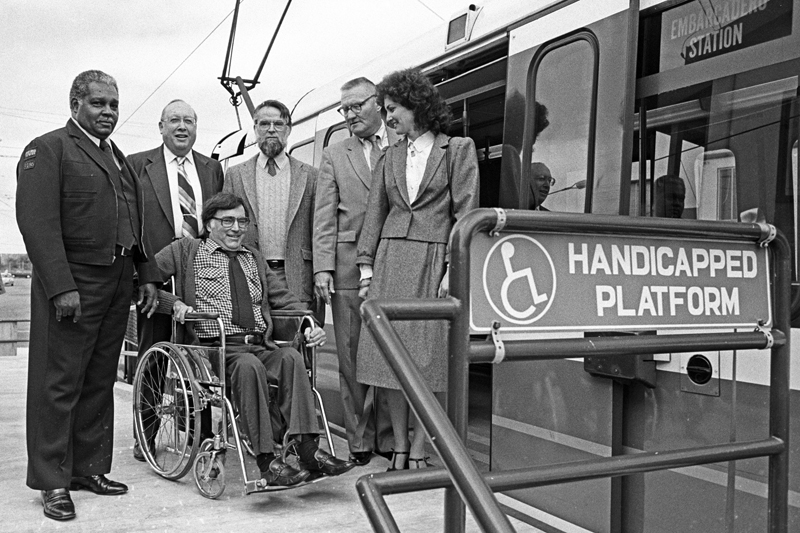 A man in a wheelchair is accompanied by a Muni operator and several others standing on a new Muni accessibility platform in 1983.