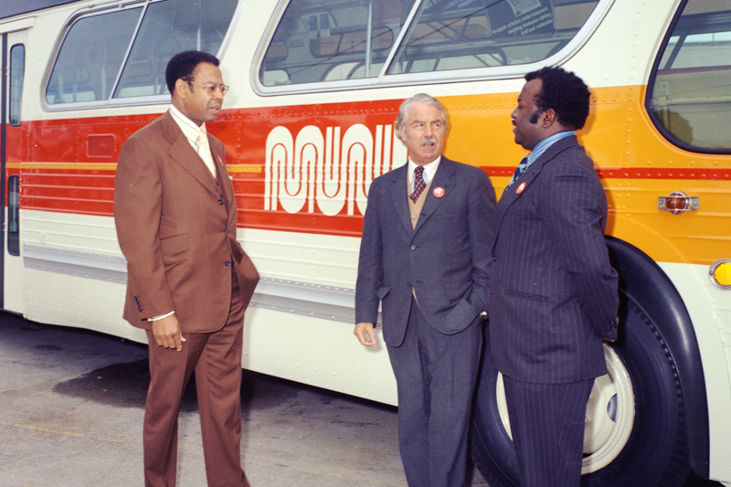 Three men standing alongside a Muni bus painted in new white, orange, and yellow colors in the 1970s