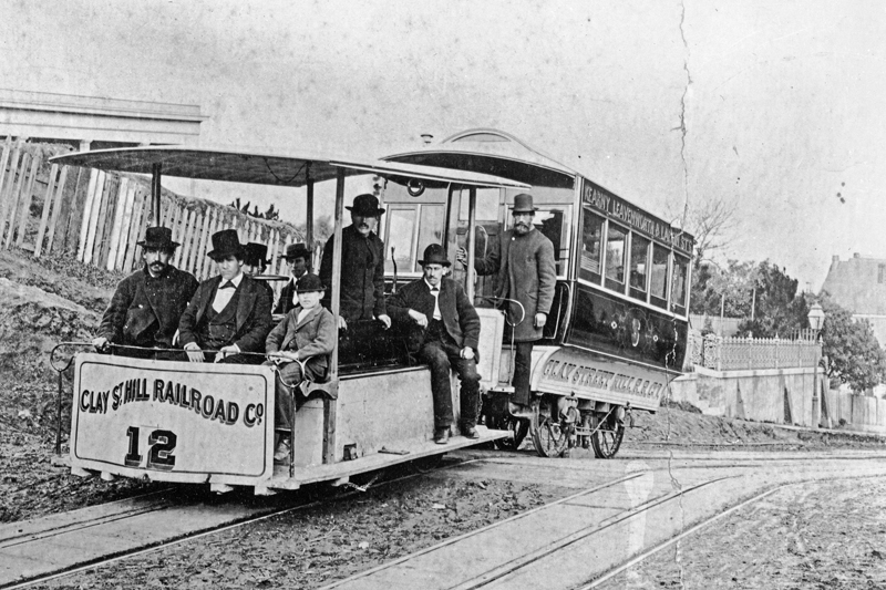 A Clay Street cable train in 1873 with people on board. An open-sided car pulls an enclosed passenger "trailer"