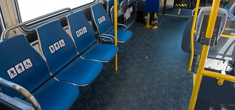 Interior of Muni coach showing blue colored priority seats near the front of the bus, seats are in a row at the left side of the image and have decals on them indicating their use.