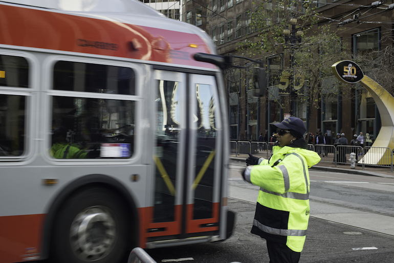Woman in cold-weather, reflective gear stands in Market Street, directing a red and gray Muni bus through the intersection with a "Super Bowl City" arched sign in the background.