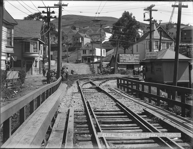 Black and white photo showing a view north on Diamond Street near Chenery in San Francisco's Glen Park Neighborhood. in the foreground is a wooden bridge and streetcar rails, followed by a street curving uphill with a handful of early 20th century wooden houses and buildings.