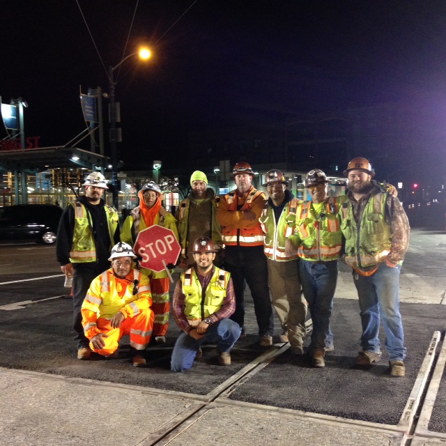 Group of construction workers stand and kneel at night with safety vests and hard hats with the Caltrain station behind them.