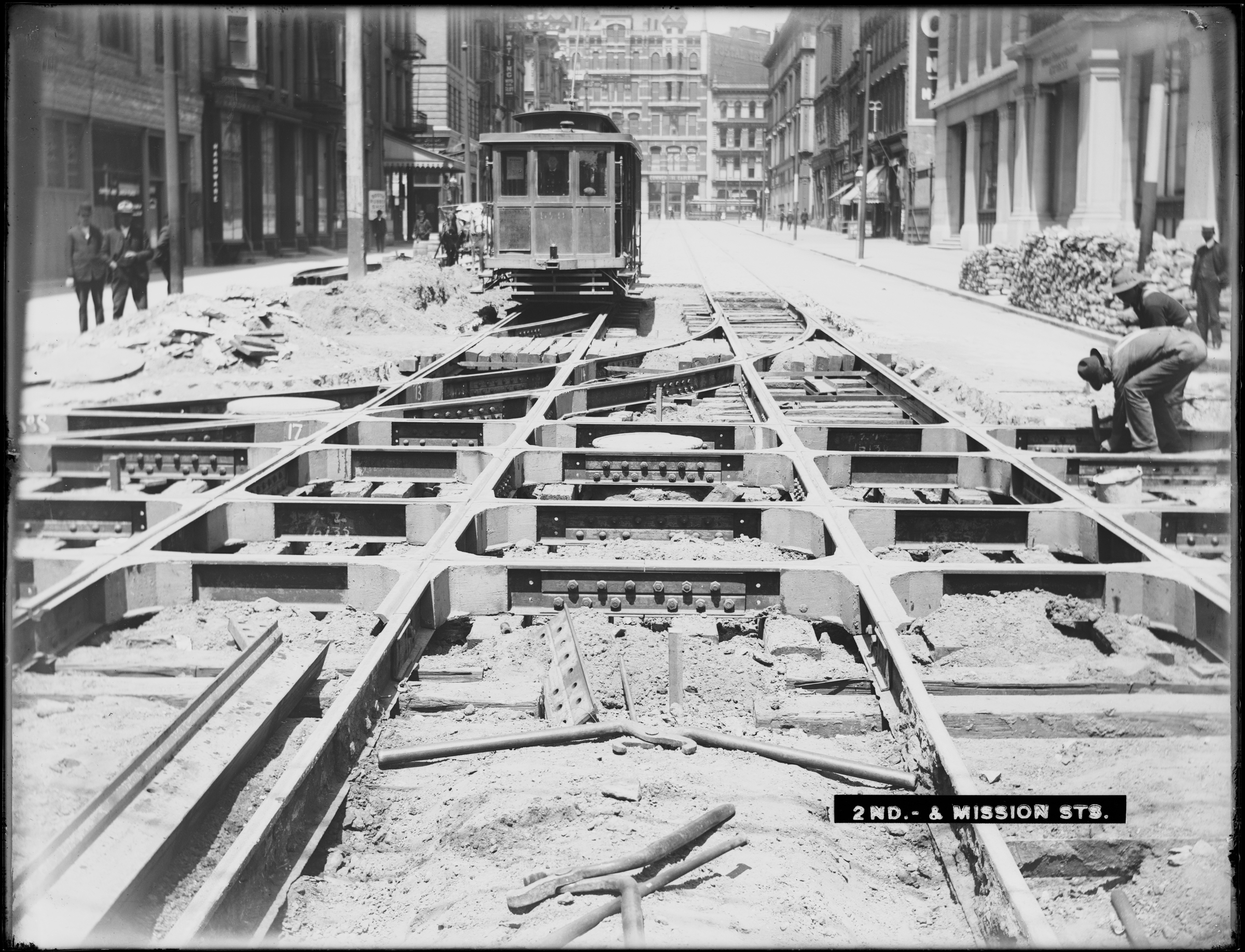 Historic black and white photograph of streetcar 578 on 2nd Street at Mission Street. The foreground is dominated by the grid of tracks still being laid. Two crewmen are working on the tracks on the right side. Two pedesri