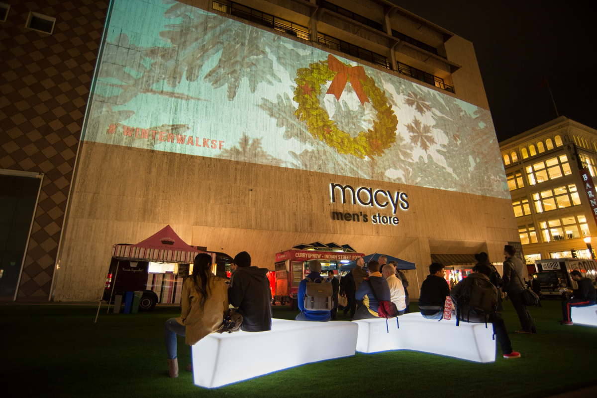 Night shot of people sitting on white, illuminated benches on astroturf with a projected image of a wreath and snowflakes on the Macy's Mens' Store building behind them.