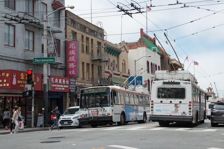 Two Muni coaches passing each other as people use a crosswalk in Chinatown during the day.
