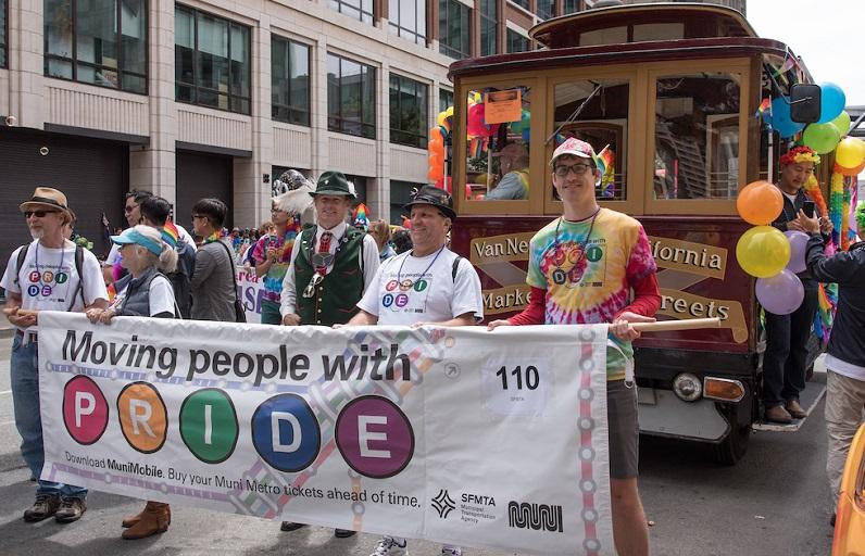 SFMTA staff in the contingent in the Pride parade, standing in front of a decorated motorized Cable Car 62 holding and a banner that says, "Moving People With Pride."