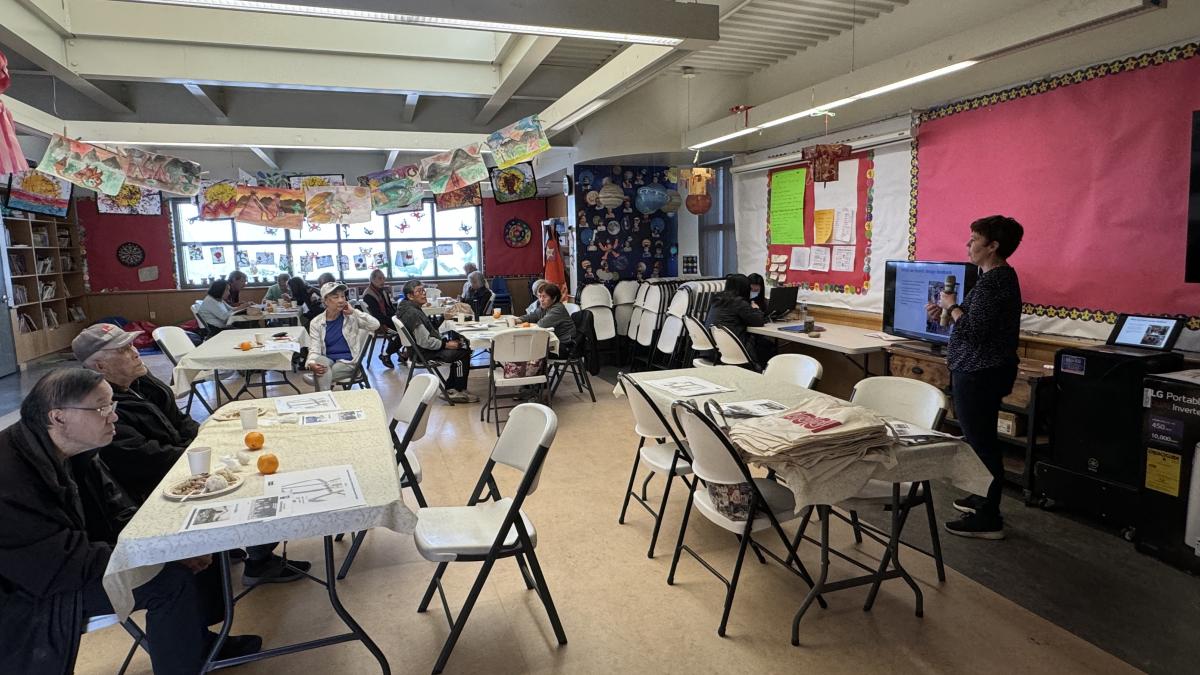 About the West Portal Rec Center, 15 people sit and eat at tables while a woman talks into a microphone and points at a map on a screen.