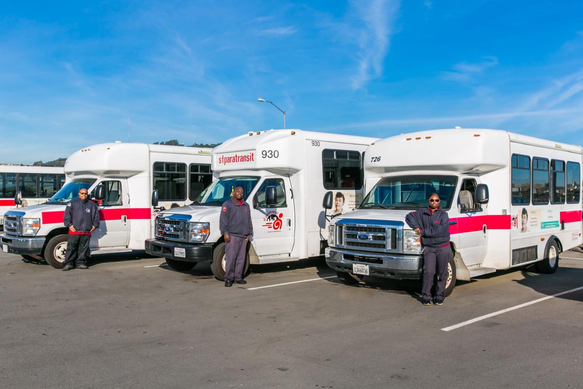 3 drivers each stand proudly in front of their paratransit vans lined up in a parking lot