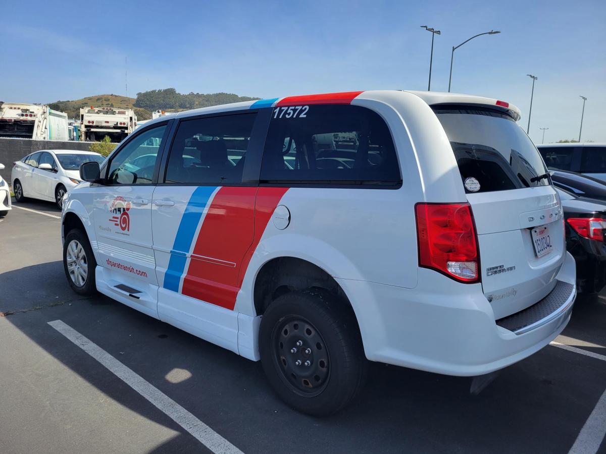 Rear three-quarter exterior view of a white Dodge Caravan SF Paratransit ramp van with red and blue diagonal striped livery parked in a parking lot