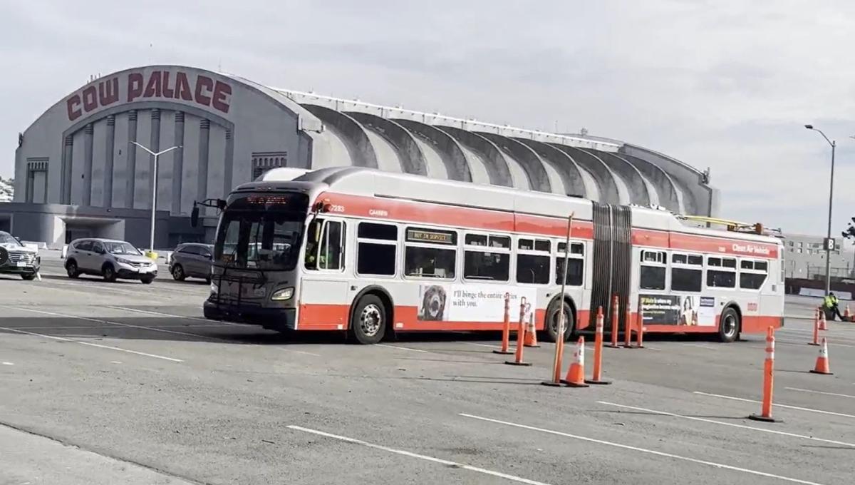 A 60-foot bus is seen in a large parking lot. A building with a curved roof is in the background. the words 'Cow Palace' appear in large red letters across the front of the building.