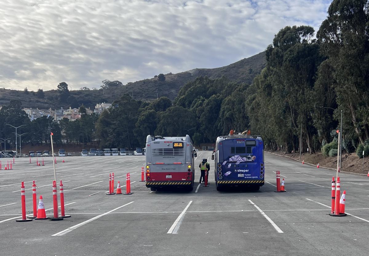 Two buses are parked side-by-side in an expansive parking lot. Orange traffic cones are visible