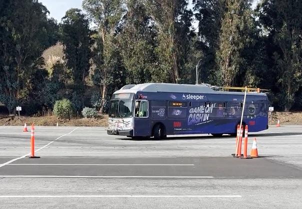 A 40-foot bus drives around a spacious parking lot. Trees are in the background. Orange traffic cones are visible in the foreground.