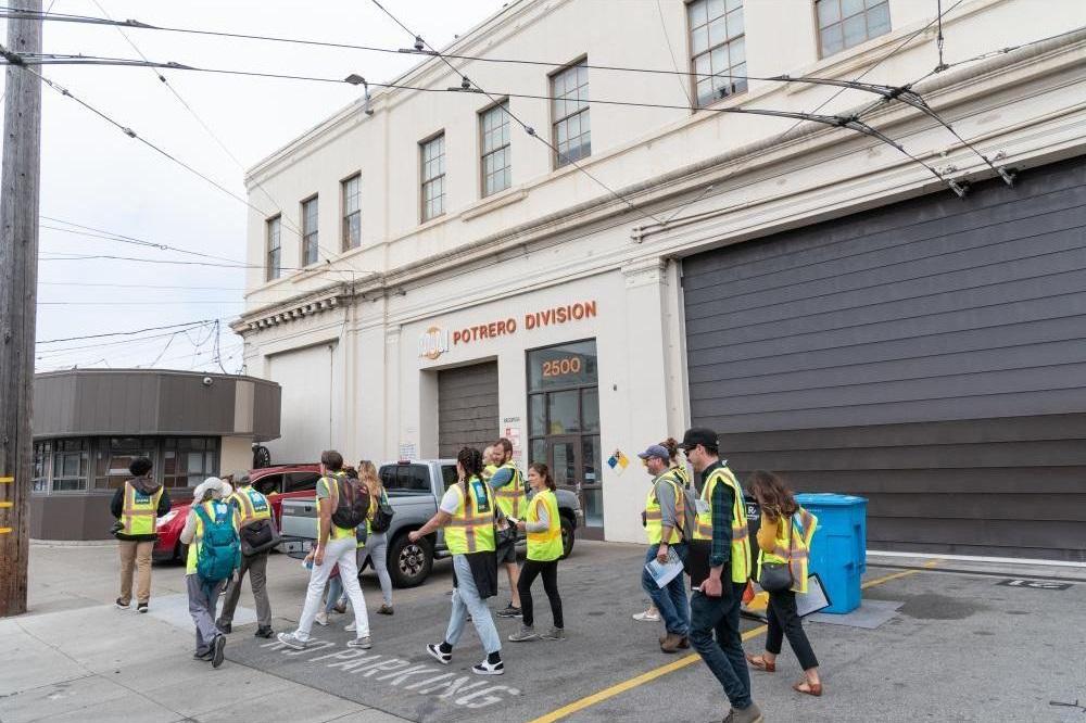 people in yellow safety vests walk along the exterior of a transit facility. A sign on the wall reads Potrero division