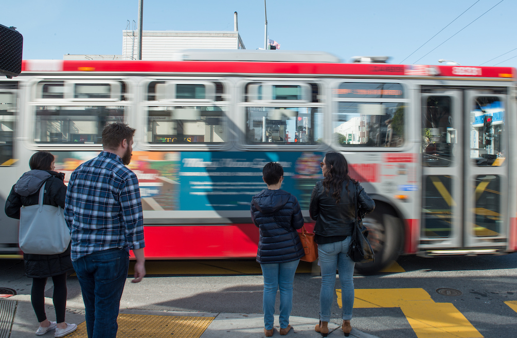 Muni bus passes corner where people are standing.