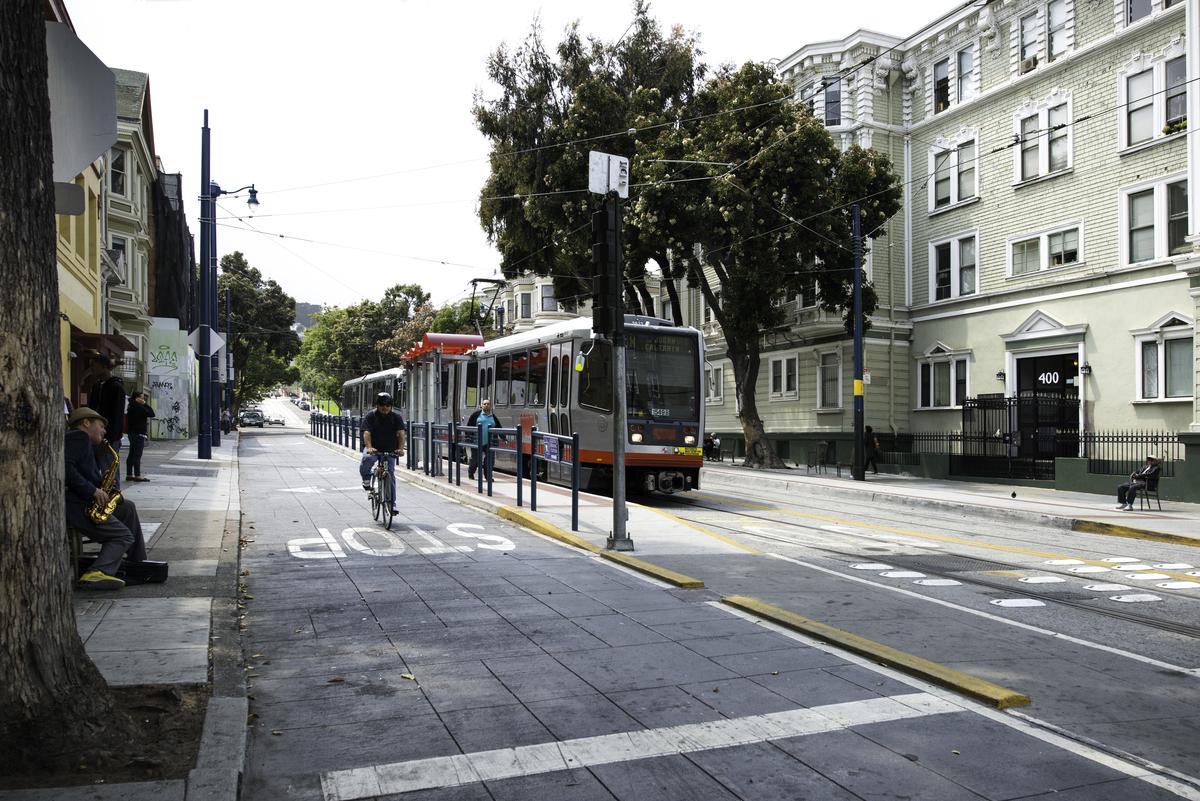 Light rail train stopped at Church and Duboce station with pedestrians and cyclist on street.