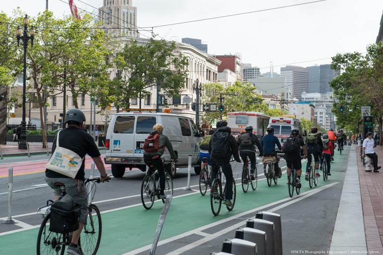 Group of cyclists riding on green protected bike lane on Market Street with vehicles and buildings in background.