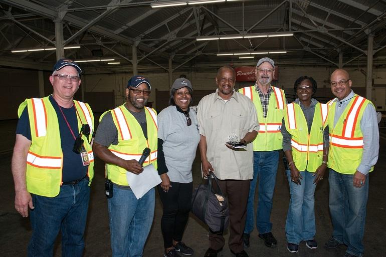 Seven transit workers in safety vests and caps stand together in an industrial warehouse setting, with Kevin Grady in the center wearing a light-colored shirt.