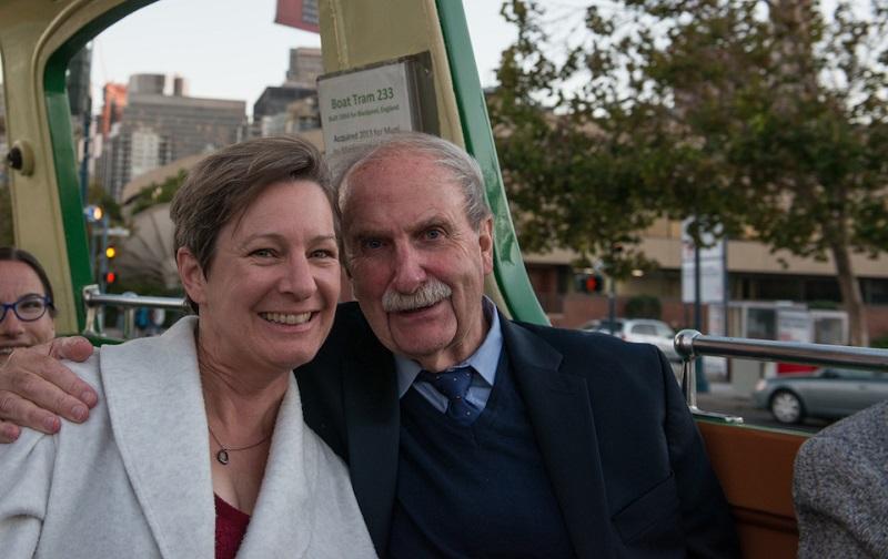 Man and woman sitting together on an open-air streetcar with city buildings visible in the background.