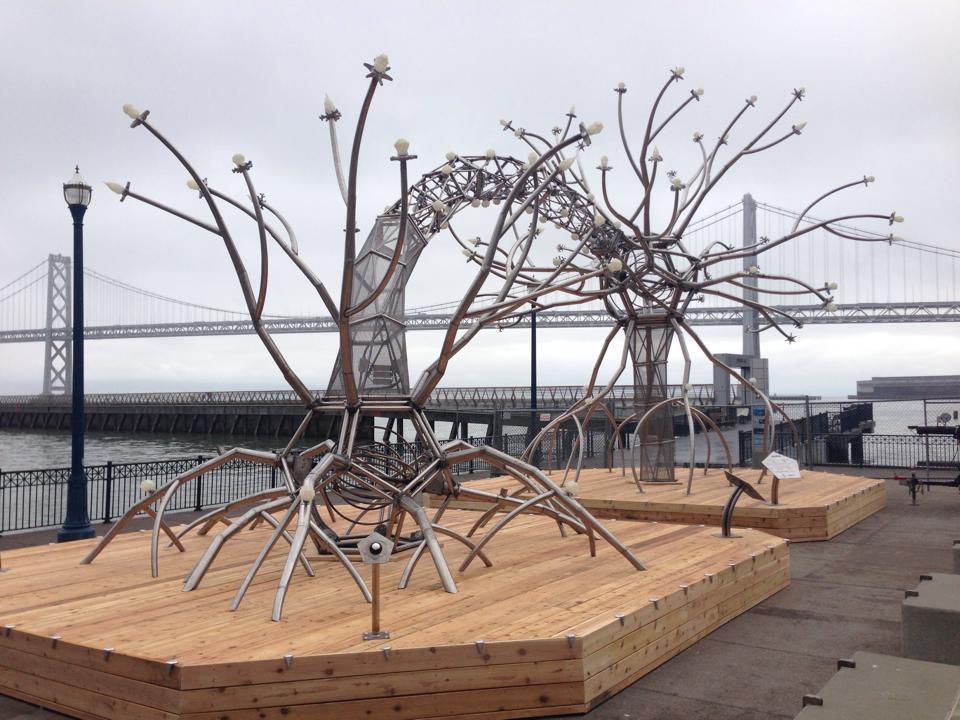 Metal tree sculpture with branching arms on wooden platform overlooking Bay Bridge.