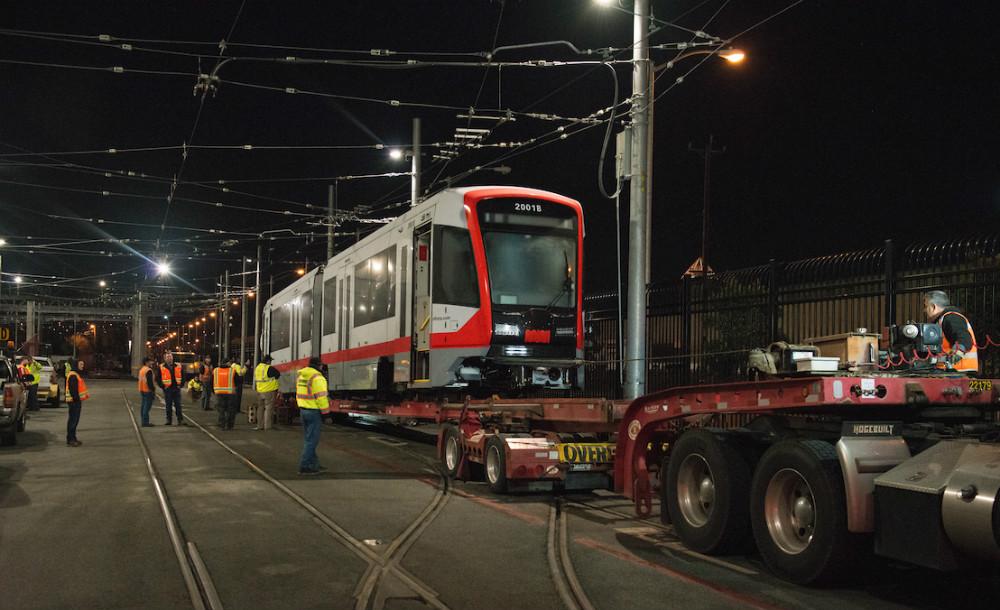 Crews offload a new red and white Muni light rail vehicle from a truck bed at night.