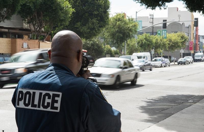 Police officer using LIDAR speed enforcement device on urban street with passing vehicles.