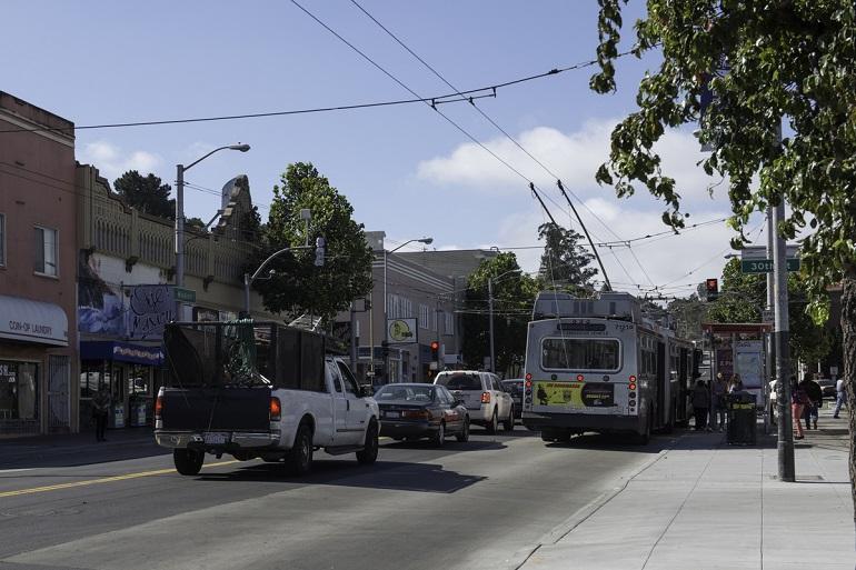 Muni trolley bus at Mission Street stop with passengers boarding, cars and trucks waiting at red light.