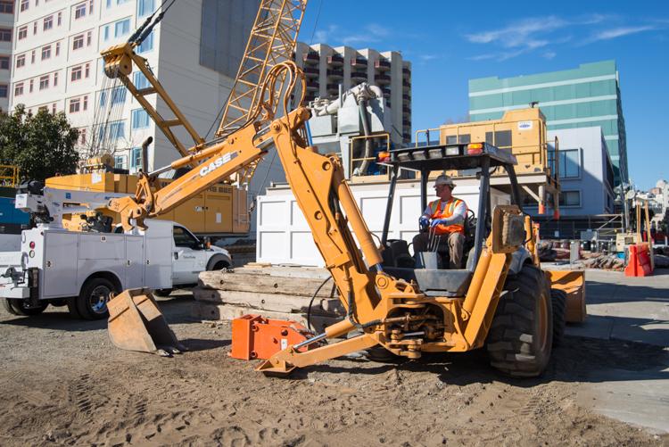 Yellow excavator with operator on sandy construction site near downtown buildings and crane