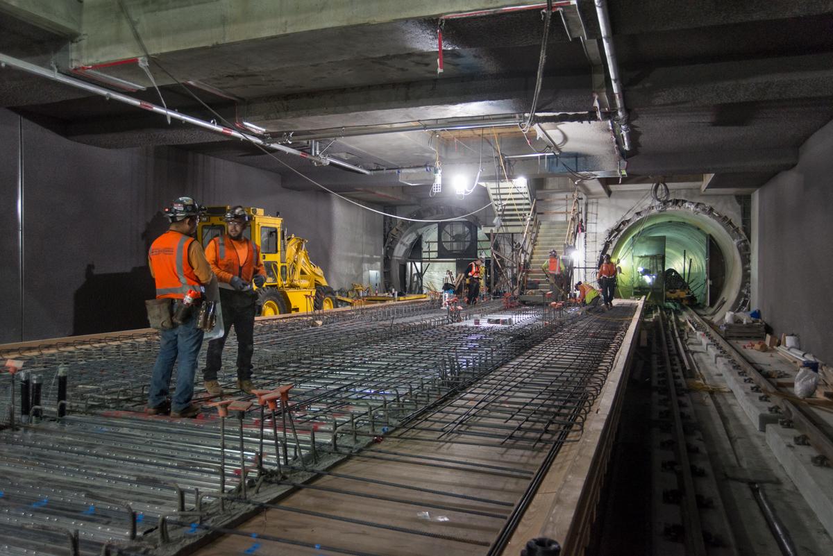 Construction workers in safety vests inspect reinforced concrete and steel framework inside an underground transit tunnel.