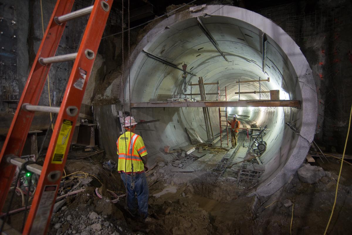 Worker in safety vest stands before large circular tunnel under construction with scaffolding and equipment visible inside.