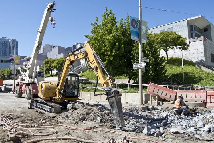 Yellow excavator demolishing concrete guide walls at 4th and Folsom streets with construction equipment and barriers visible.