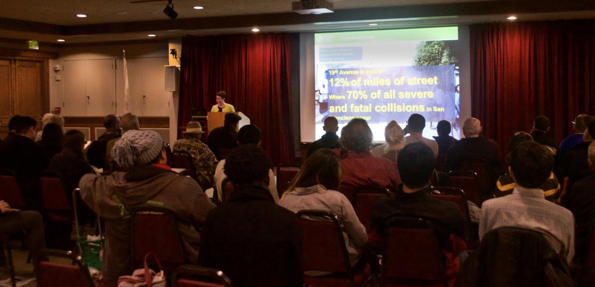 Audience seated in auditorium watching presenter at podium with projection screen displaying street safety statistics.