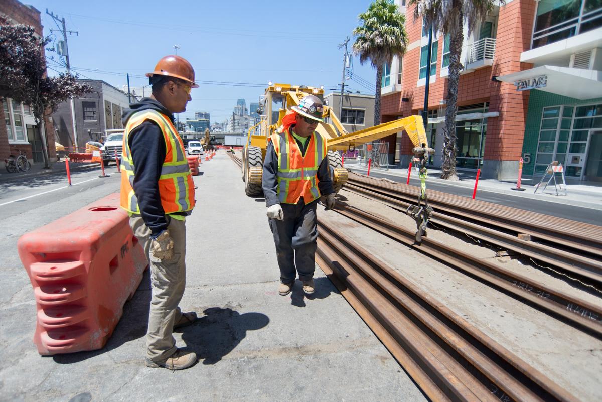 Two construction workers in safety vests inspect rail sections on a city street during infrastructure work.