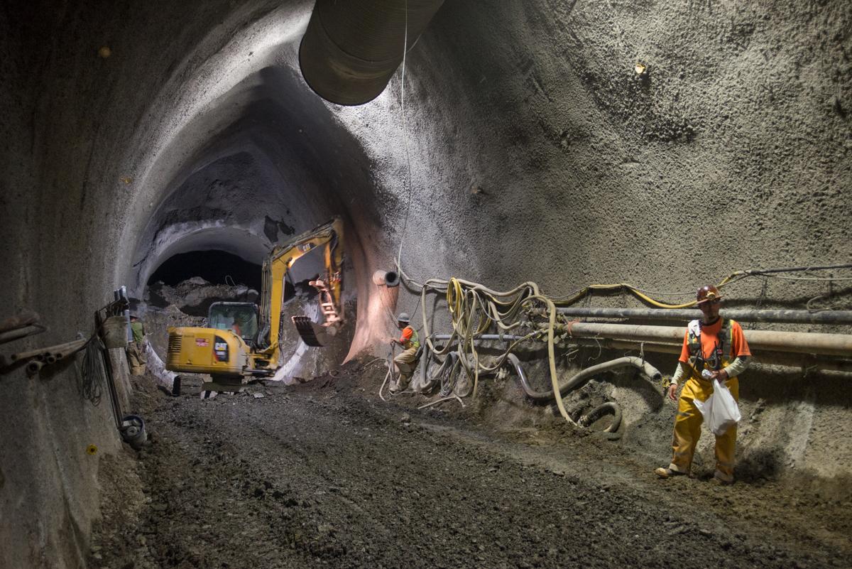 Workers in safety gear operate an excavator inside a large concrete tunnel under construction.