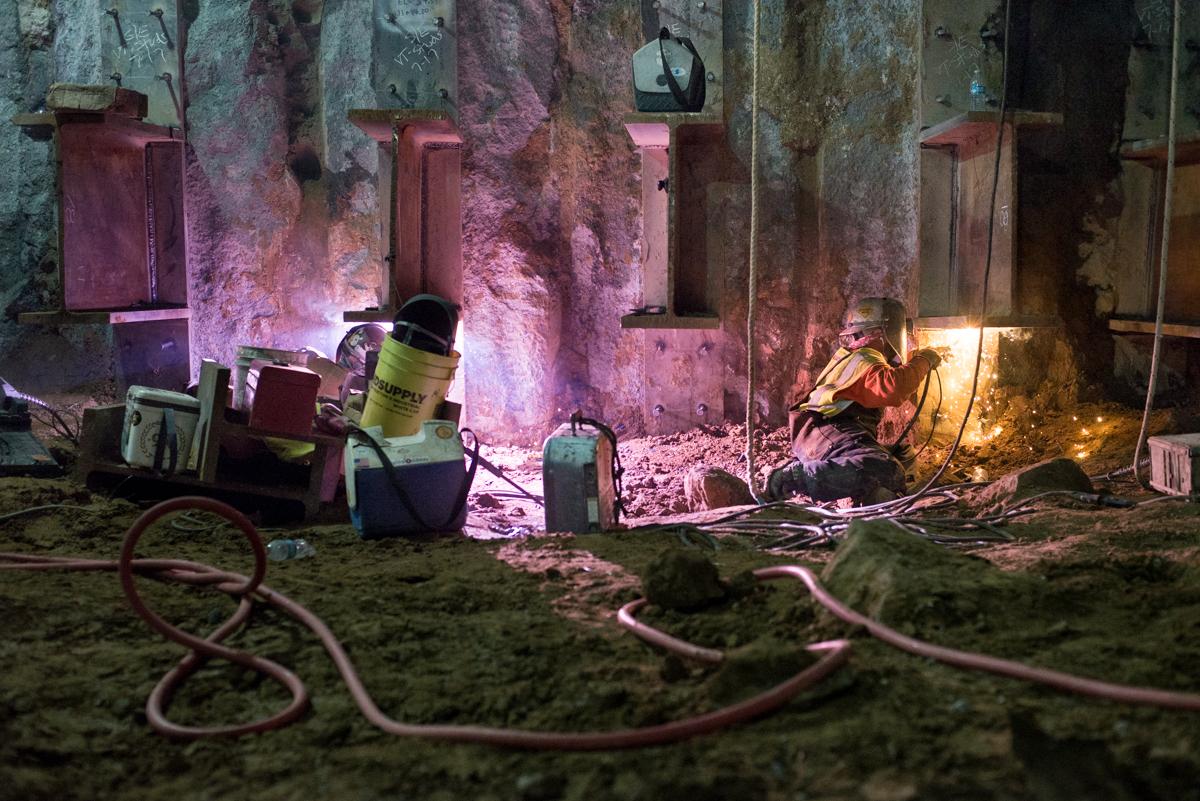 Workers weld steel I-beams to station box wall at night under pink lighting.