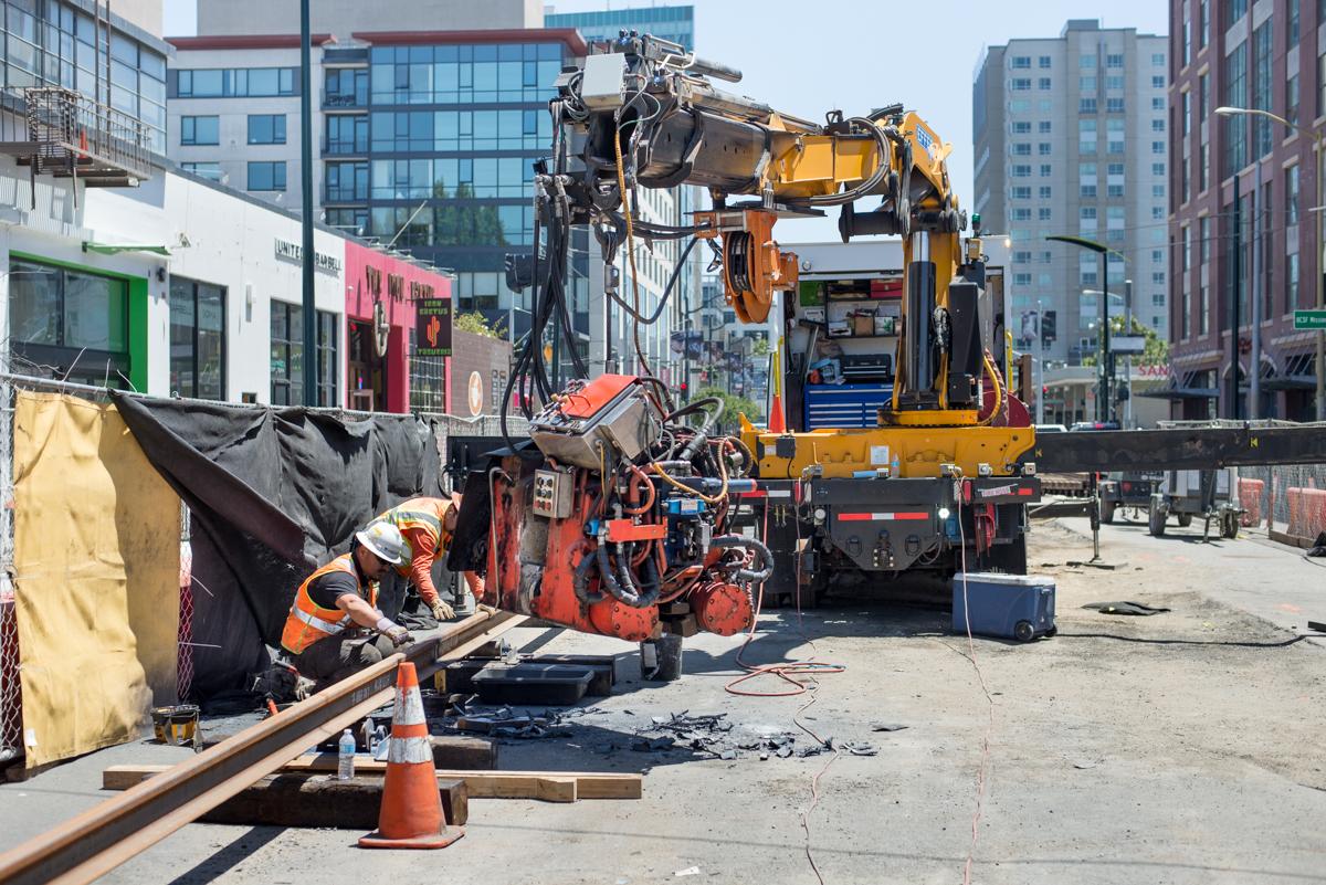 Construction worker operating rail welding equipment on urban street with crane and equipment nearby.
