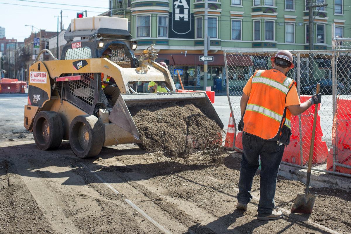 Construction worker in orange vest operates skid steer loader spreading gravel at street work site.