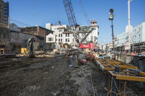 Construction site with excavation equipment and cranes at Chinatown Station, surrounded by urban buildings and temporary barriers.