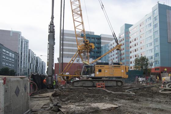 Construction crew operates yellow drilling equipment at underground transit station site surrounded by urban buildings.