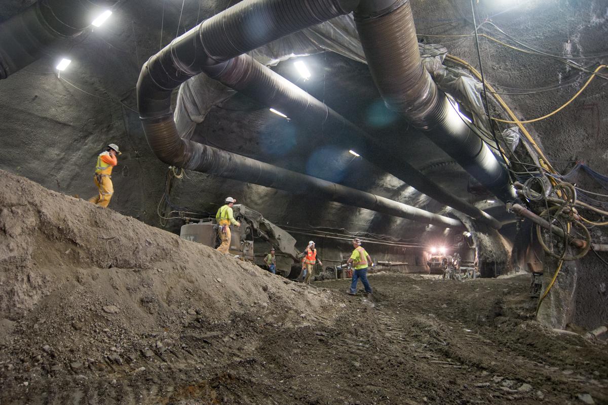 Workers in safety gear inside underground tunnel with large pipes and construction equipment overhead.