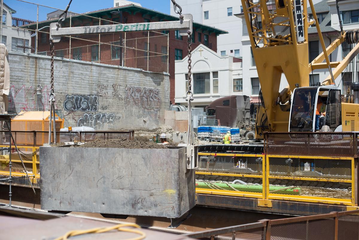 Construction equipment and excavation bucket at underground transit station entrance with surrounding buildings.