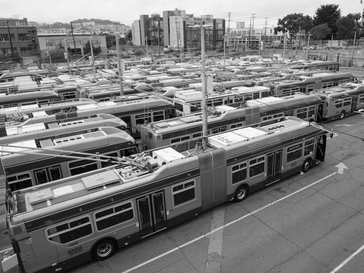 Members from the public and a tour guide wearing an orange hat are seen on the upper level of the Potrero Yard, overlooking buses parked below.