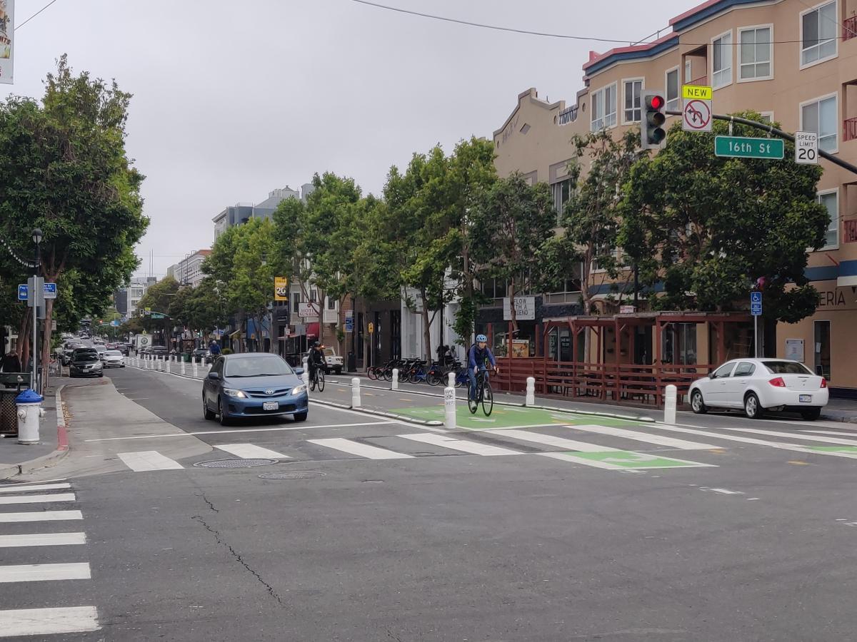 Image shows the intersection of Valencia and 16th Street, with a cyclist in the center-running bikeline, a car waiting at the light, cars parked along the curb, and a parklet.